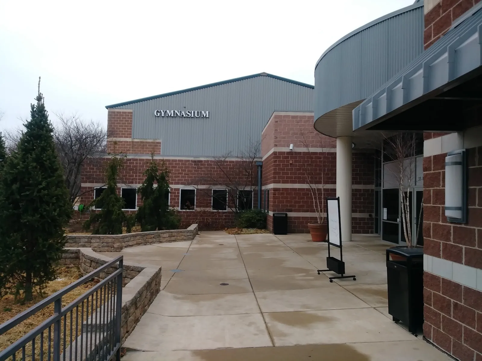 The brick and glass entrance of the Webster Groves Ice Arena, a curved brick wall beside a canopy and a pine tree under a pale sky. — Webster Groves Ice Arena