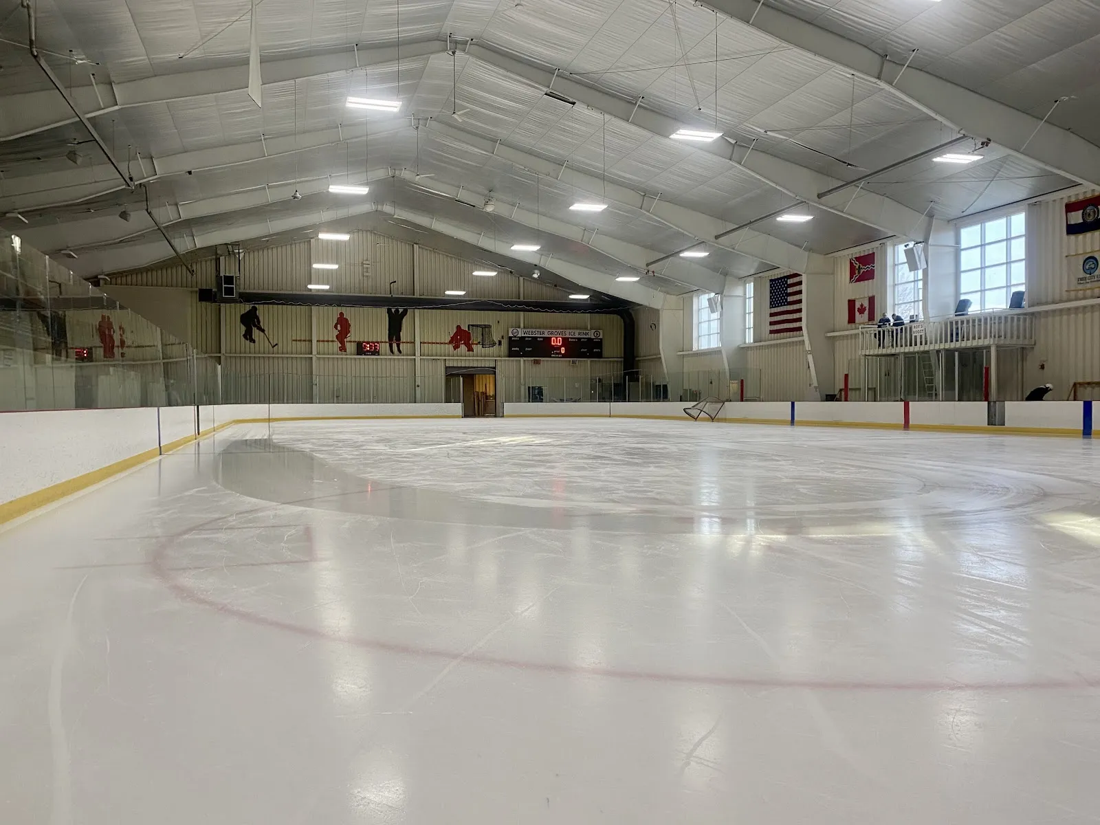 Empty ice sheet with red/blue face-off lines and a hanging scoreboard reading 'WEBSTER GROVES ICE RINK'; American, Canadian and Missouri flags visible on a mezzanine above the boards. — Webster Groves Ice Arena
