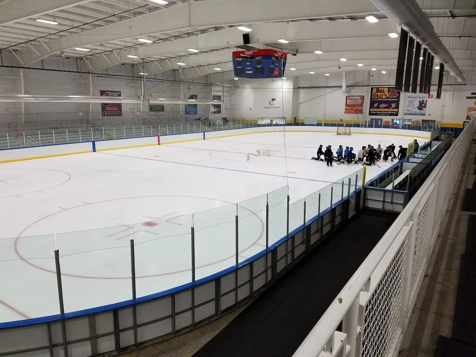 Una pista de Webster Ice Arena desde el pasillo superior, con un equipo reunido junto a las vallas del fondo bajo un techo blanco y el marcador — Webster Ice Arena