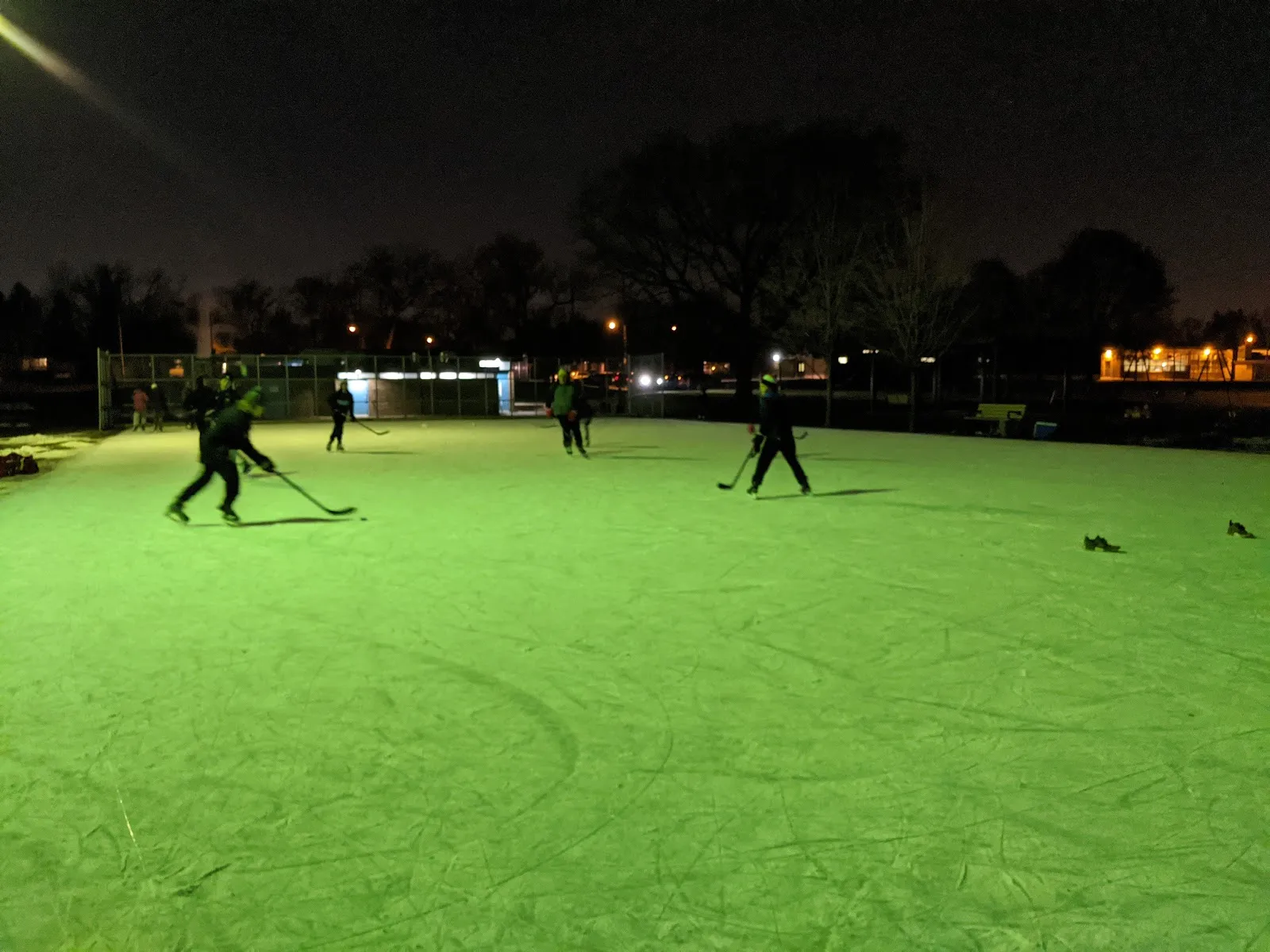 Hockey being played at night on the Wedgewood Park rink under floodlights, with the park and trees behind the boards — Wedgewood Park - Etobicoke Outdoor Ice Rink