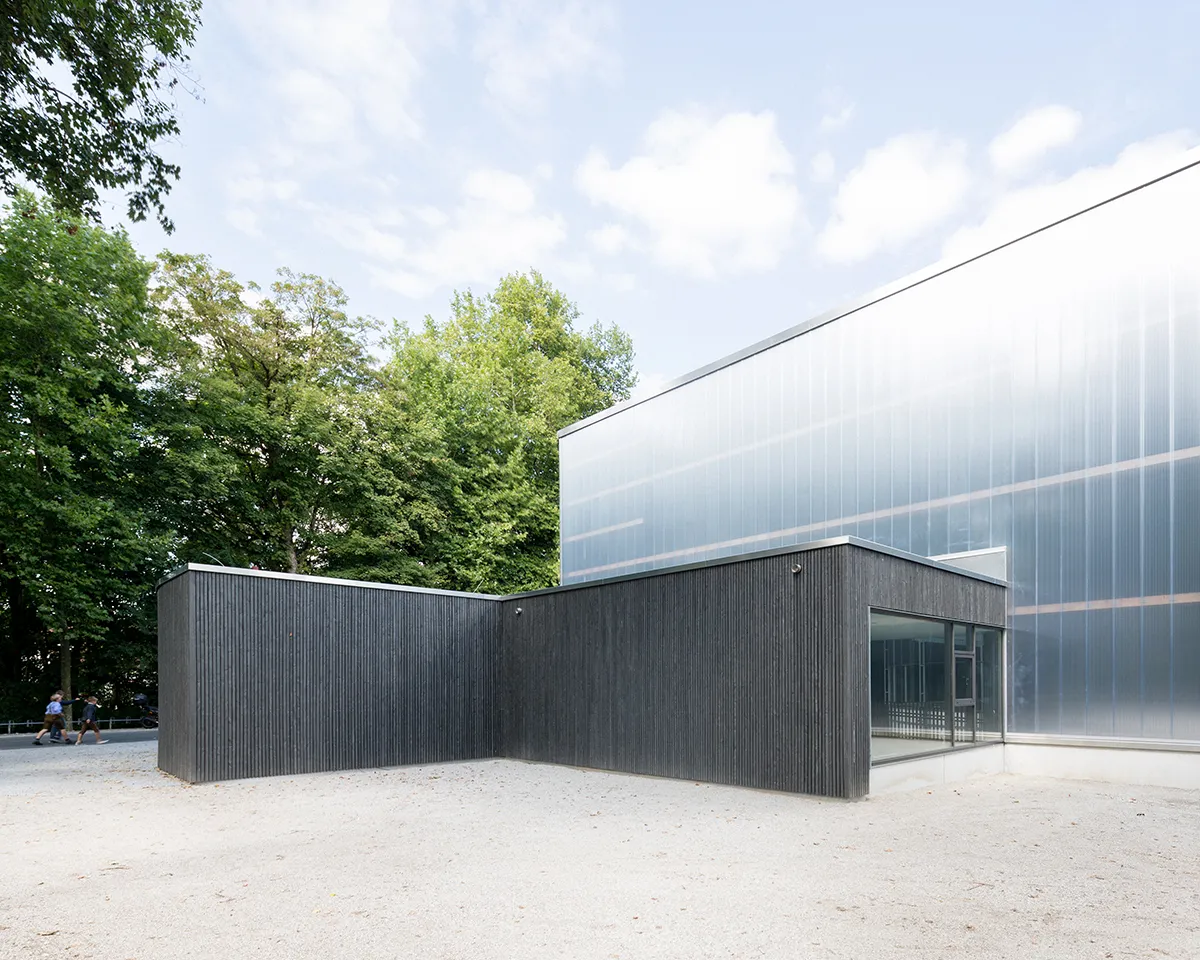 Der durchscheinende obere Baukörper der Halle über einem niedrigen, schwarz holzverkleideten Eingangsblock, zwischen Bäumen und einem Kiesvorplatz bei Tageslicht. – Weihenstephan Arena (Eishalle Freising)
