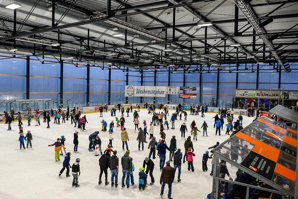 Ein gut besuchter Publikumslauf auf dem Eis der Weihenstephan Arena in Freising, mit dem Stahldach, der blauen Wand und der Weihenstephan-Bandenwerbung hinter den Läufern. – Weihenstephan Arena (Eishalle Freising)