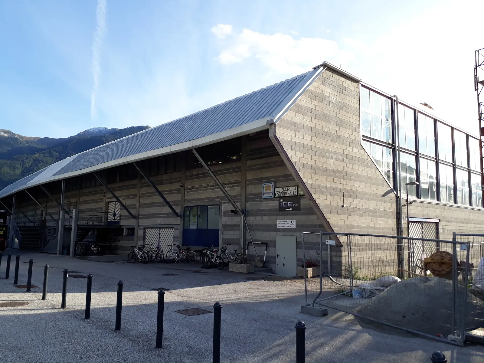 Il fianco della Weihenstephan Arena di Vipiteno, una parete rivestita in pietra sotto un lungo tetto metallico spiovente, con le montagne sullo sfondo – Weihenstephan Arena
