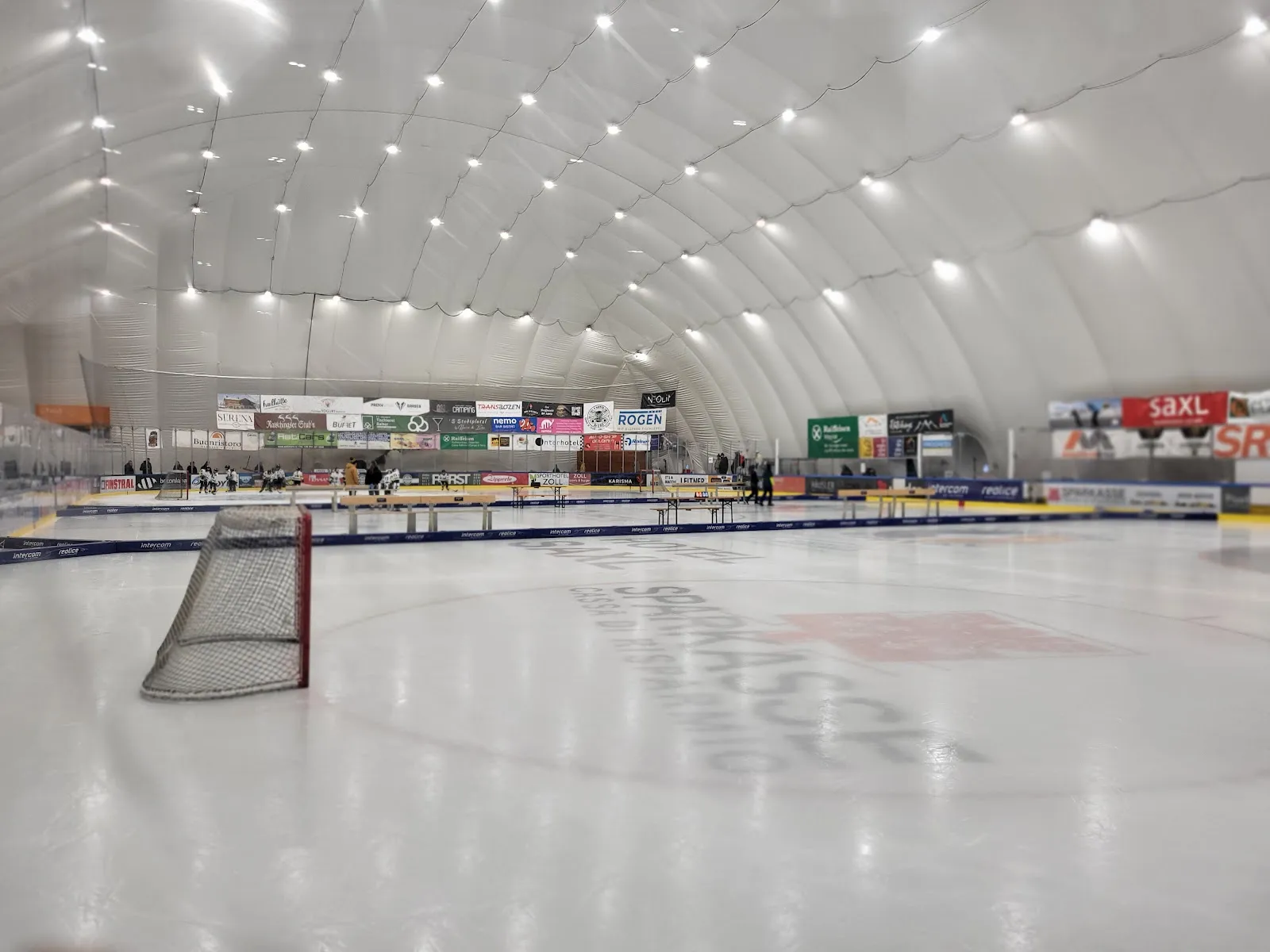 Il ghiaccio sotto la cupola bianca pressostatica della Weihenstephan Arena di Vipiteno, con una porta a sinistra e alcune persone lontane lungo le balaustre – Weihenstephan Arena