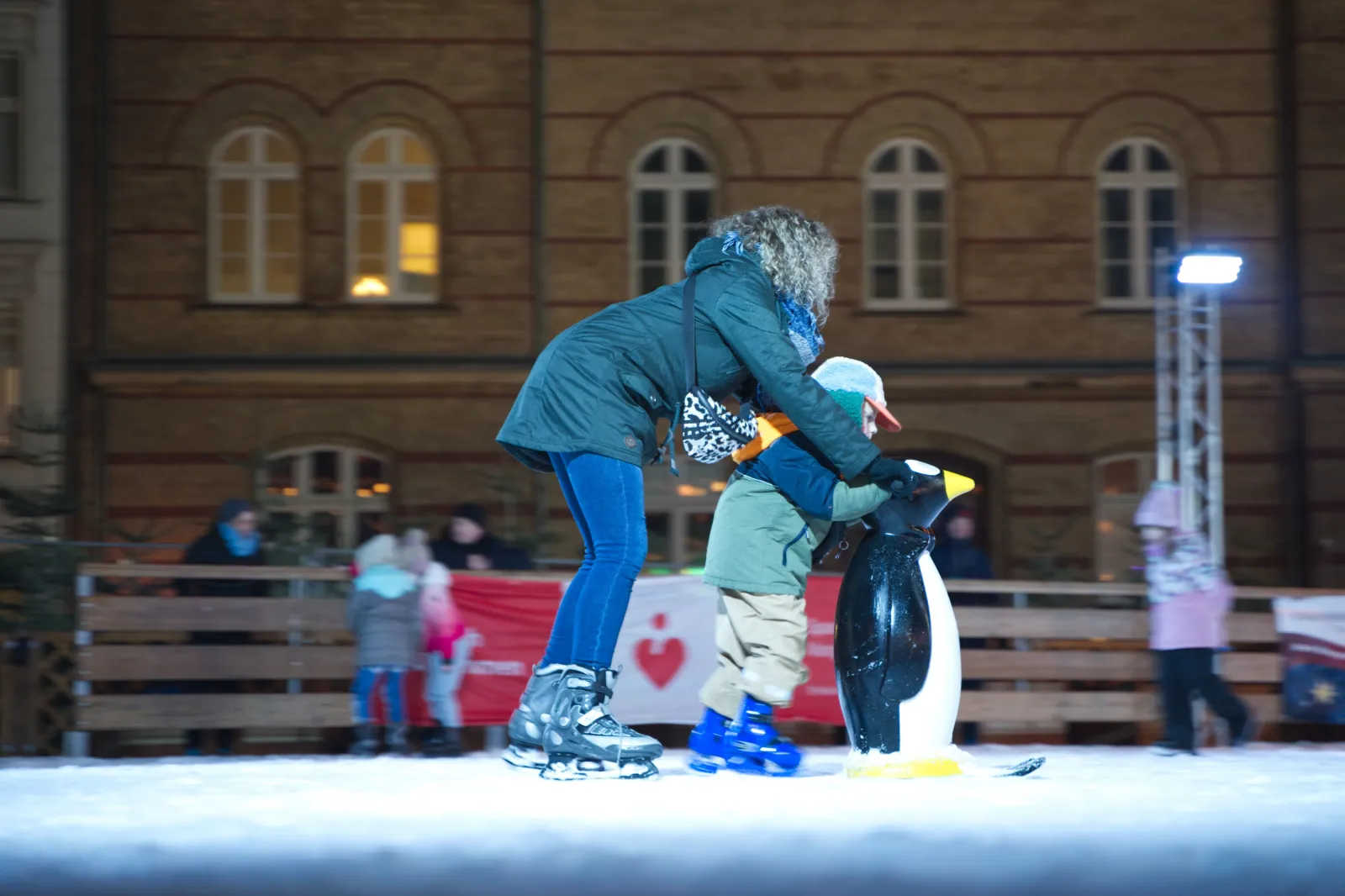 An evening skating session on the Neuer Markt rink: an adult in a teal jacket helps a small child on skates push a black-and-white penguin skating aid across the ice, with wooden boards, a red banner and a brick façade behind. — Eisbahn Stralsunder Weihnachtsmarkt