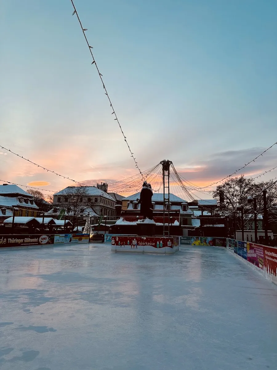 The empty ice on the Theaterplatz at dusk, with the wooden Christmas-market huts and snowy roofs behind the boards under a pink and blue sky. — Weimar on Ice (Theaterplatz)
