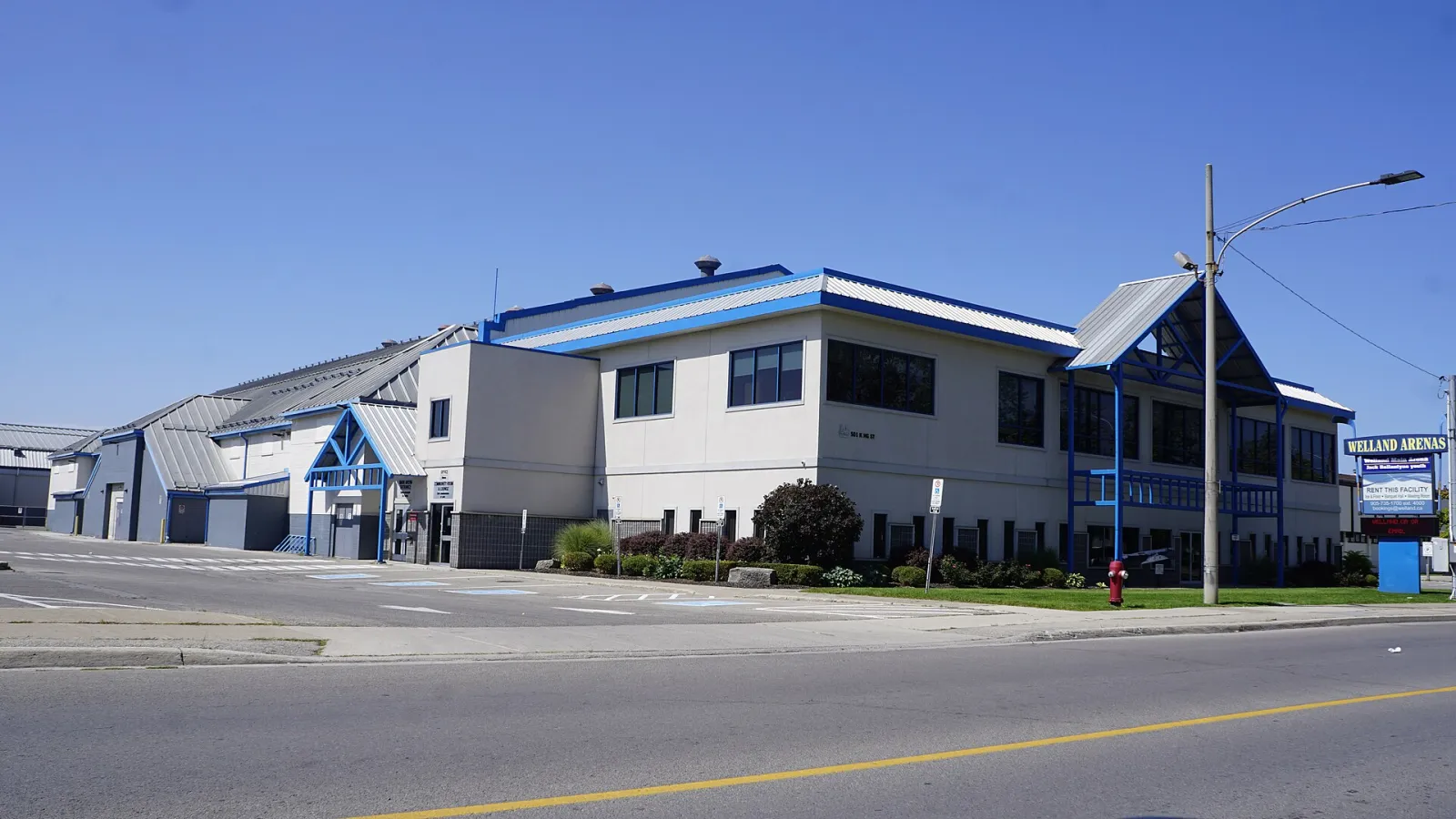 El edificio blanco y azul de Welland Arenas en la esquina de King Street, con su letrero WELLAND ARENAS junto a la calle y estacionamiento al lado. — Welland Arenas (LIV Communities & Jack Ballantyne)