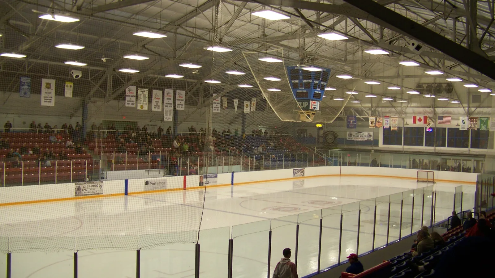 El hielo vacío de la Welland Main Arena desde los asientos, con gradas rojas y azules, y estandartes y banderas colgados de las armaduras de acero del techo. — Welland Arenas (LIV Communities & Jack Ballantyne)
