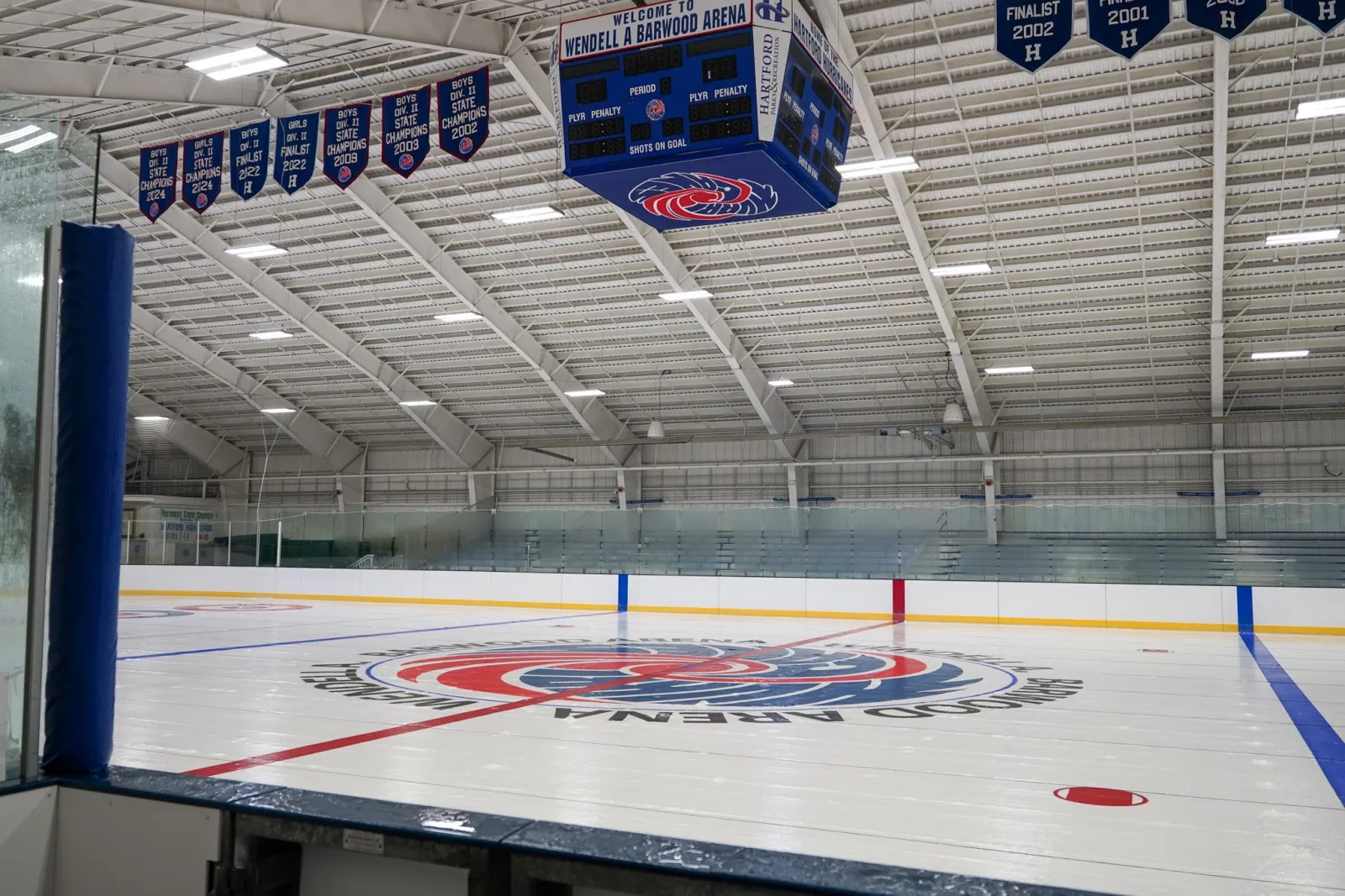 La pista vacía desde las vallas de una esquina, con el escudo de la arena en el centro de la pista, un marcador central colgante y estandartes de campeonatos bajo un techo blanco. — Wendell A. Barwood Arena