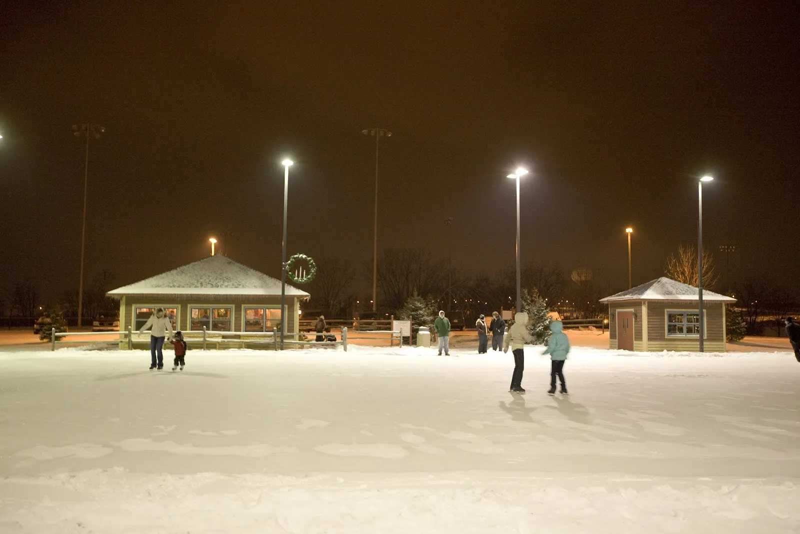 Evening skating on the outdoor ice at Wentworth Park, with two small park buildings and tall lights beyond the rink. — Wentworth Ice Rink
