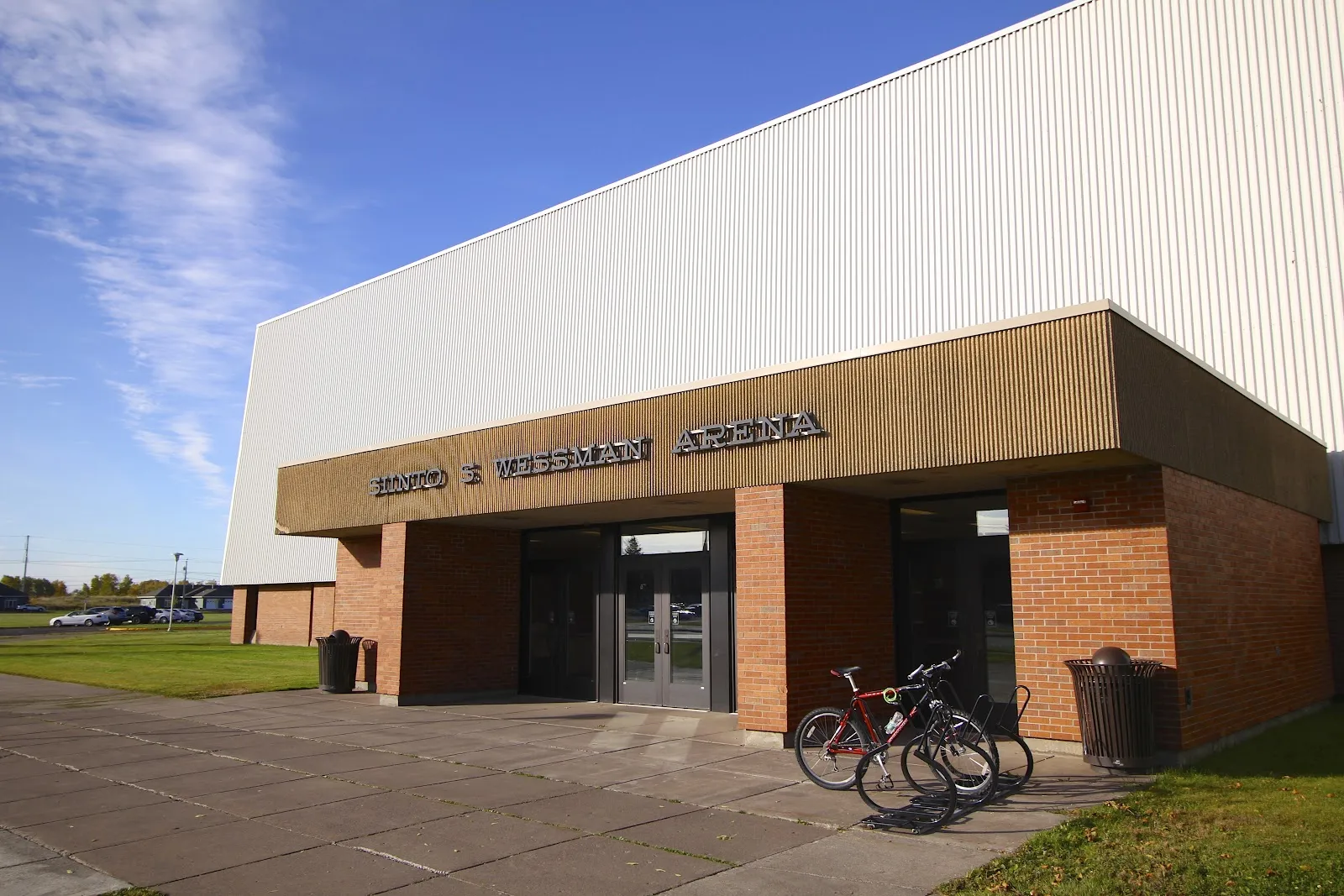 The entrance of the Siinto S. Wessman Arena, with its name in raised letters over brick piers and glass doors, bikes at a rack and the tall metal-sided arena behind. — Wessman Arena