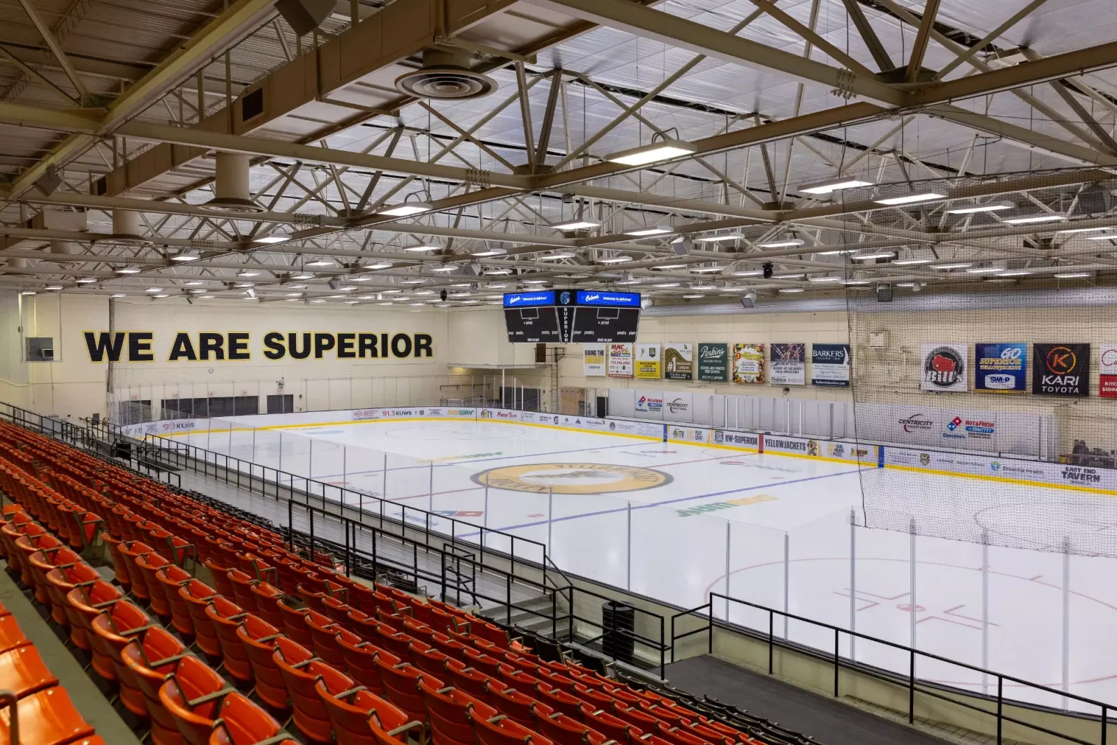 The empty sheet seen from the orange seats, with 'We Are Superior' on the far wall and the center-hung scoreboard under the steel trusses. — Wessman Arena