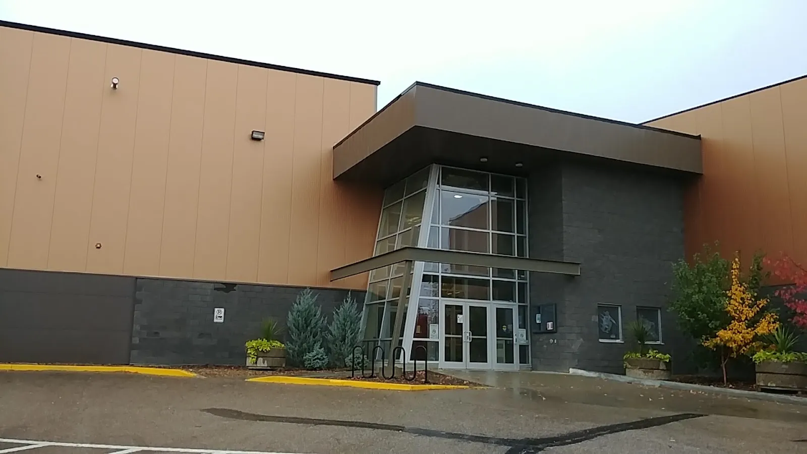 A glass-fronted entrance of the West Central Events Centre between the tall tan wall panels of its arena boxes, with planters and a bike rack by the doors — West Central Events Centre