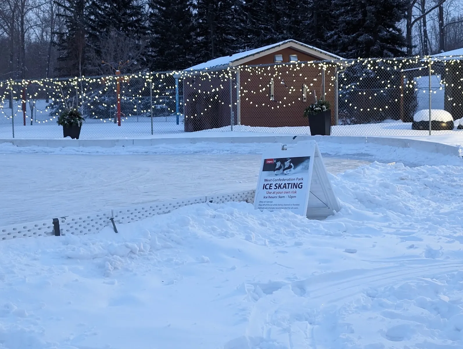 La pista al aire libre de West Confederation Park bajo la nieve, con sus series de luces, el refugio para calentarse y el letrero de patinaje sobre hielo, sin personas a la vista — West Confederation Park Outdoor Skating Rink