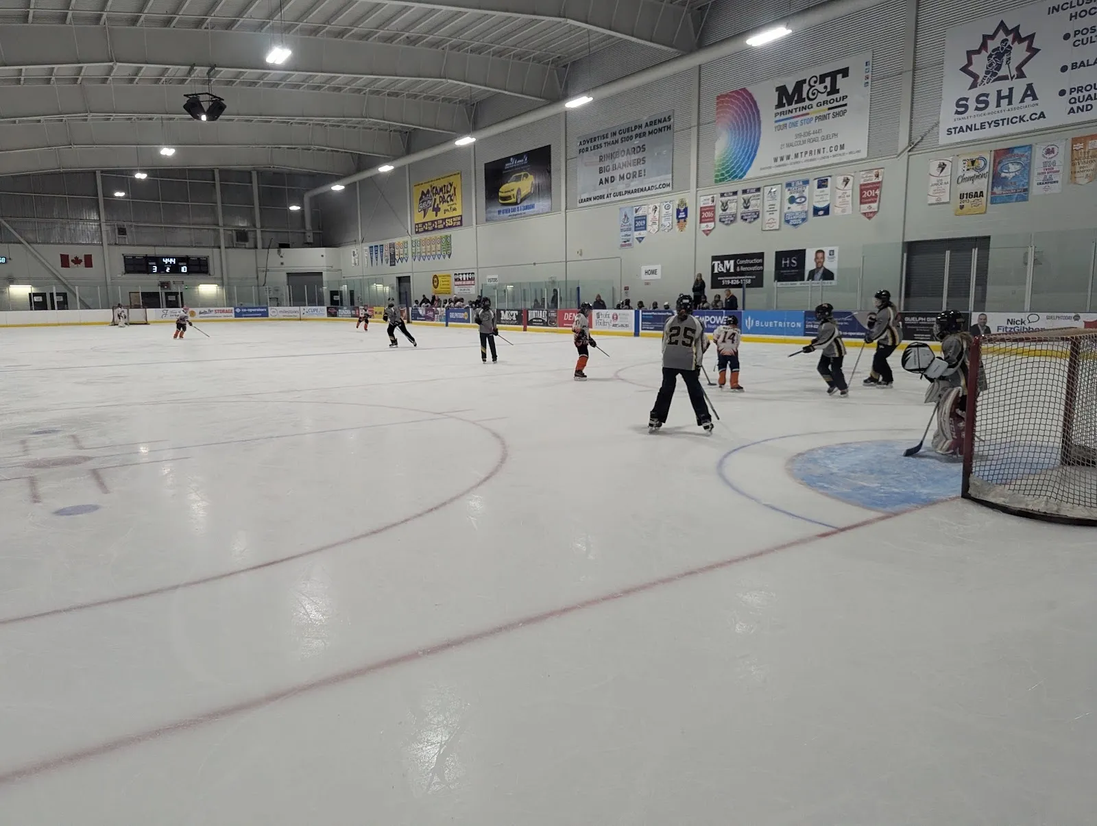 A practice on a wide sheet, players spread across the ice under a long ceiling with sponsor boards and banners along the far wall. — West End Community Centre (Guelph)