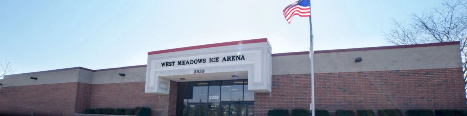 Front entrance of West Meadows Ice Arena, a brick building with red trim, an illuminated entrance sign reading WEST MEADOWS ICE ARENA above the door, and a flagpole out front — West Meadows Ice Arena