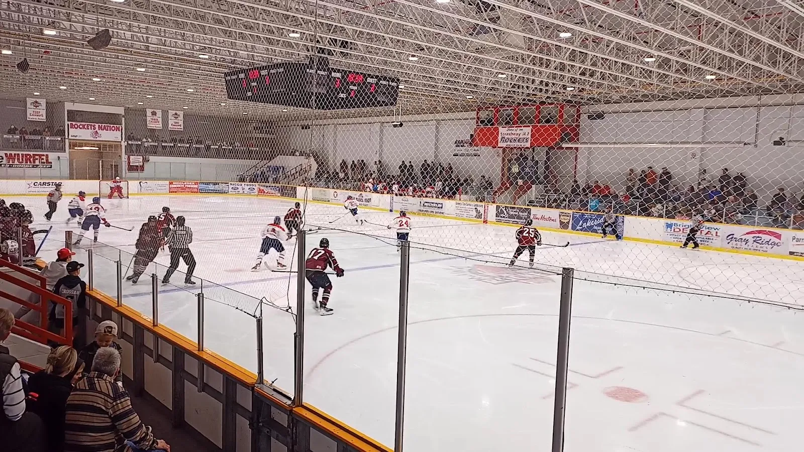 A game seen across a wide sheet through a clear rail, players spread across the ice under a steel ceiling with red scoreboards at the far end. — West Middlesex Memorial Centre (Strathroy)