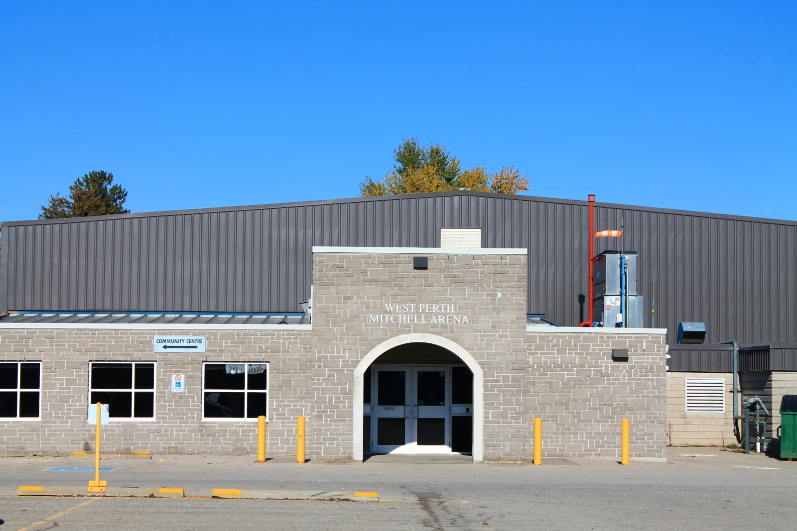 Mitchell arena entrance: a grey brick front block with an arched double door under the West Perth Mitchell Arena lettering, in front of a long dark metal-clad rink building. — West Perth Arena & Community Centre