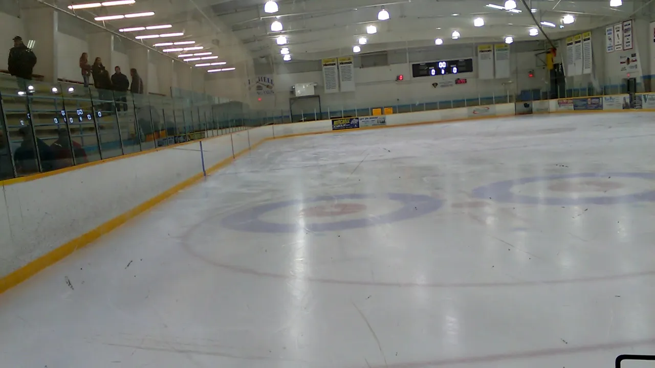 A wide empty sheet seen along the boards, grey-white ice under a long pale ceiling with a blue-lit far wall. — West Perth Arena & Community Centre
