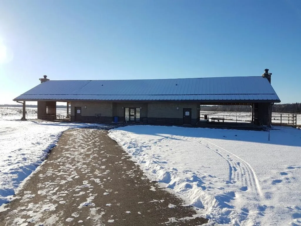 The West River's Edge pavilion beside the skating loop, a long low building with a metal roof and a trodden path through the snow. — West River's Edge (WRE) Skating Loop