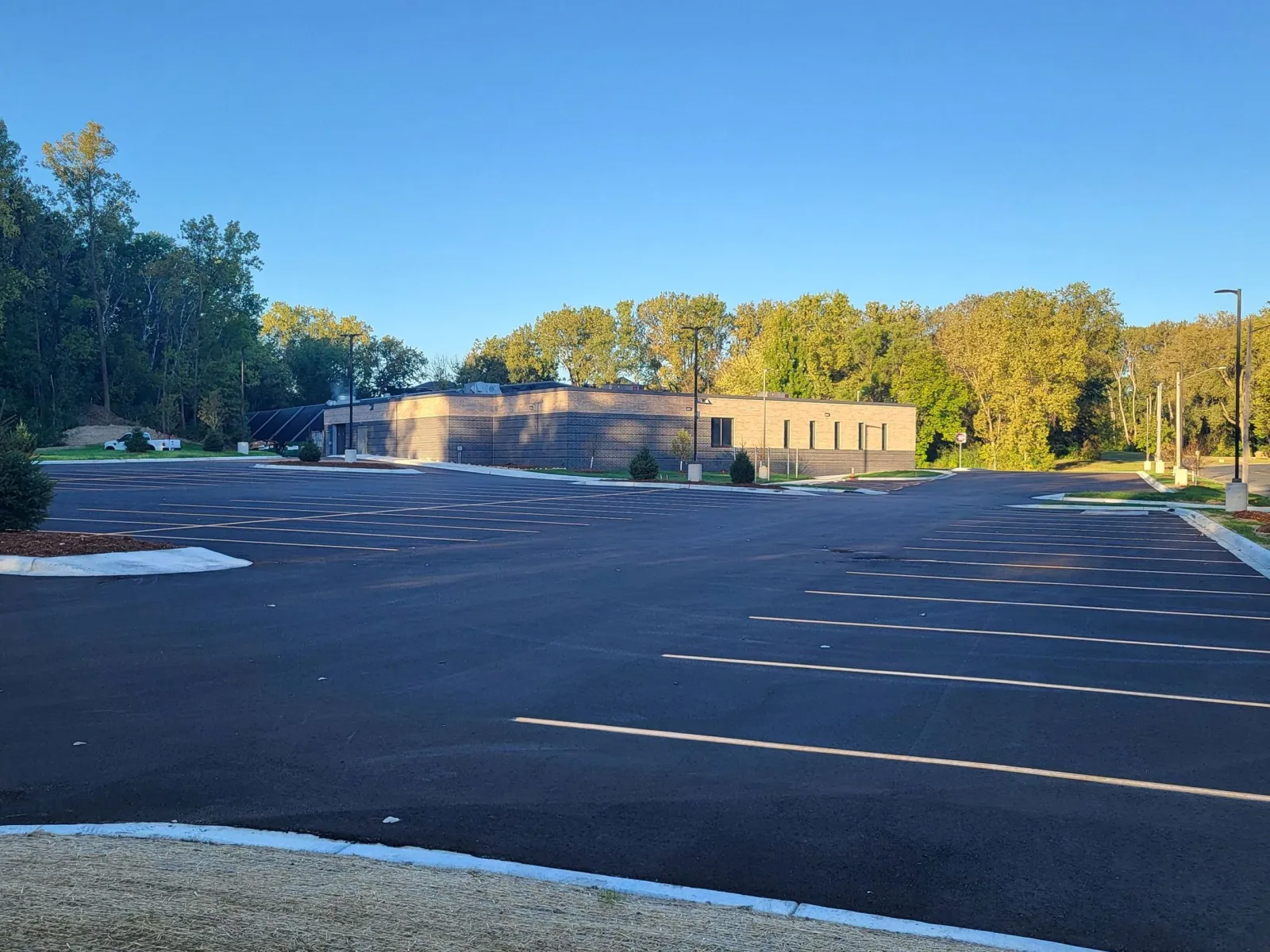 El edificio bajo de la arena, color arena y café, al fondo de su estacionamiento recién pavimentado y pintado, bajo árboles en un día despejado. — West St. Paul Ice Arena