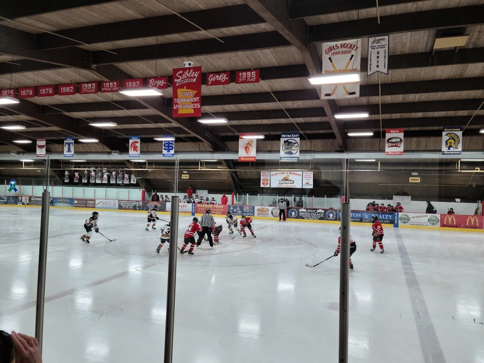 Un partido visto a través de los postes de vidrio, con jugadores repartidos por el hielo bajo un techo oscuro del que cuelgan banderines de equipos en la pared del fondo. — West St. Paul Ice Arena