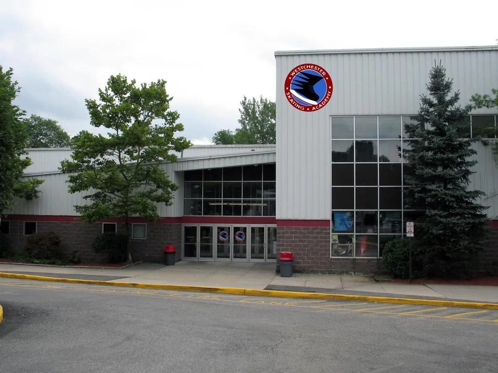 Front of a wide grey metal-clad building with a maroon brick base — Westchester Skating Academy