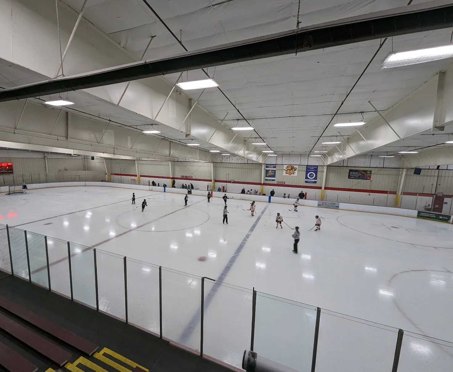 A rink at Westchester Skating Academy seen from the stands, a youth game spread across the ice under a white ceiling and team banners — Westchester Skating Academy