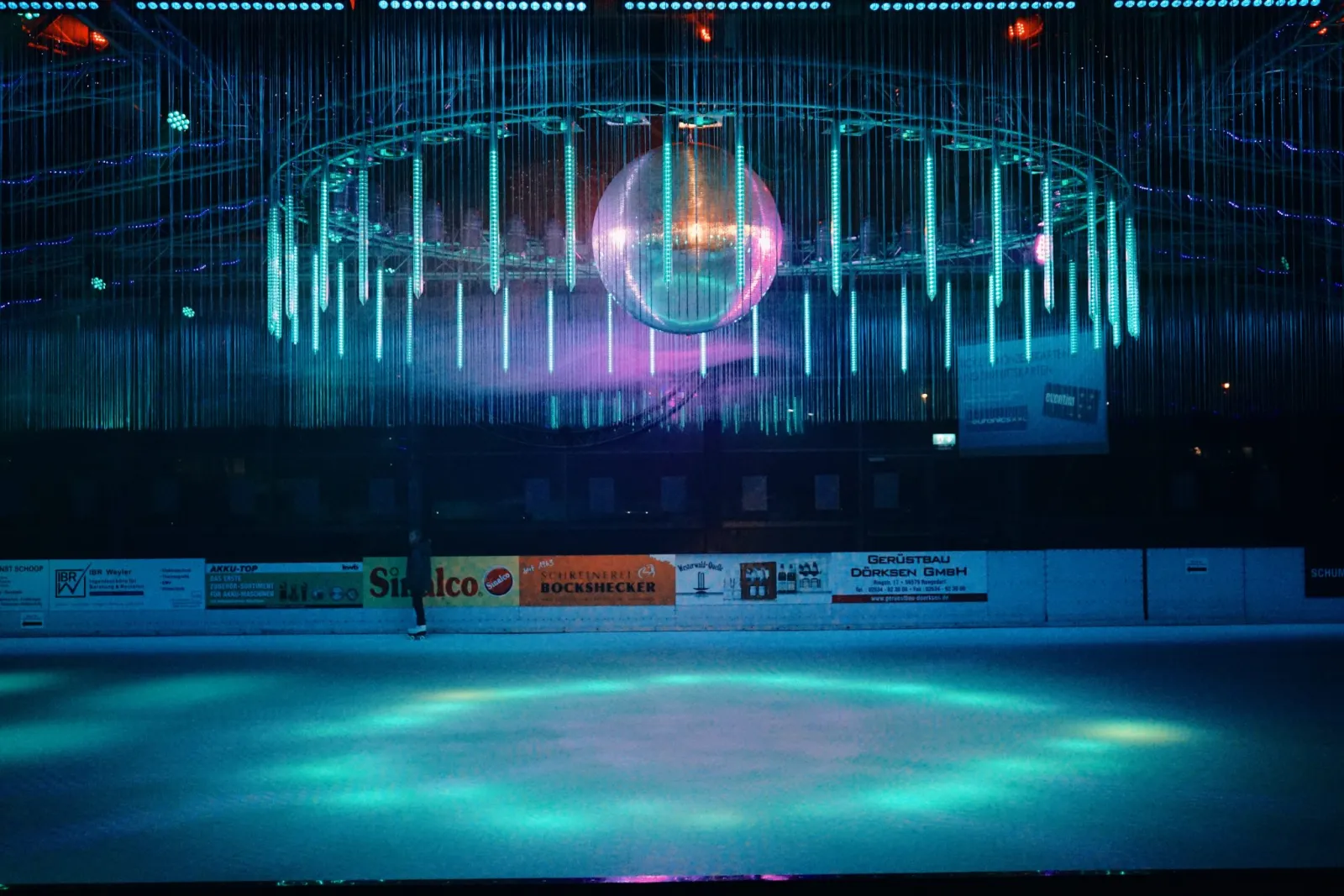 The synthetic skating floor under a ring of blue and pink lights with a mirror ball, almost empty. — Westerwald Arena Asbach (Gleit- und Rollschuhbahn)