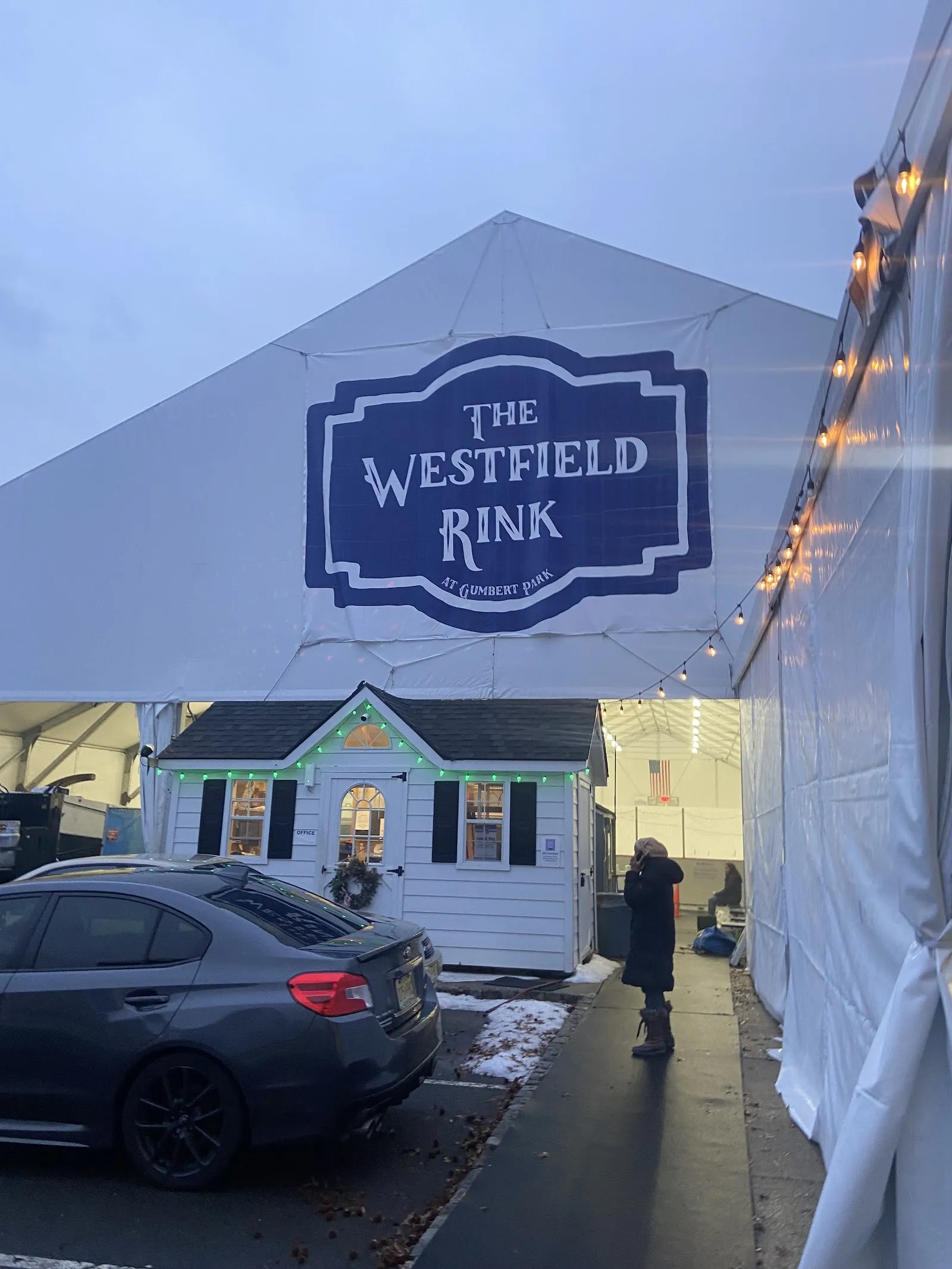The white tent over The Westfield Rink with its large name sign and the small cottage at the entrance, seen at dusk from the car park — The Westfield Rink