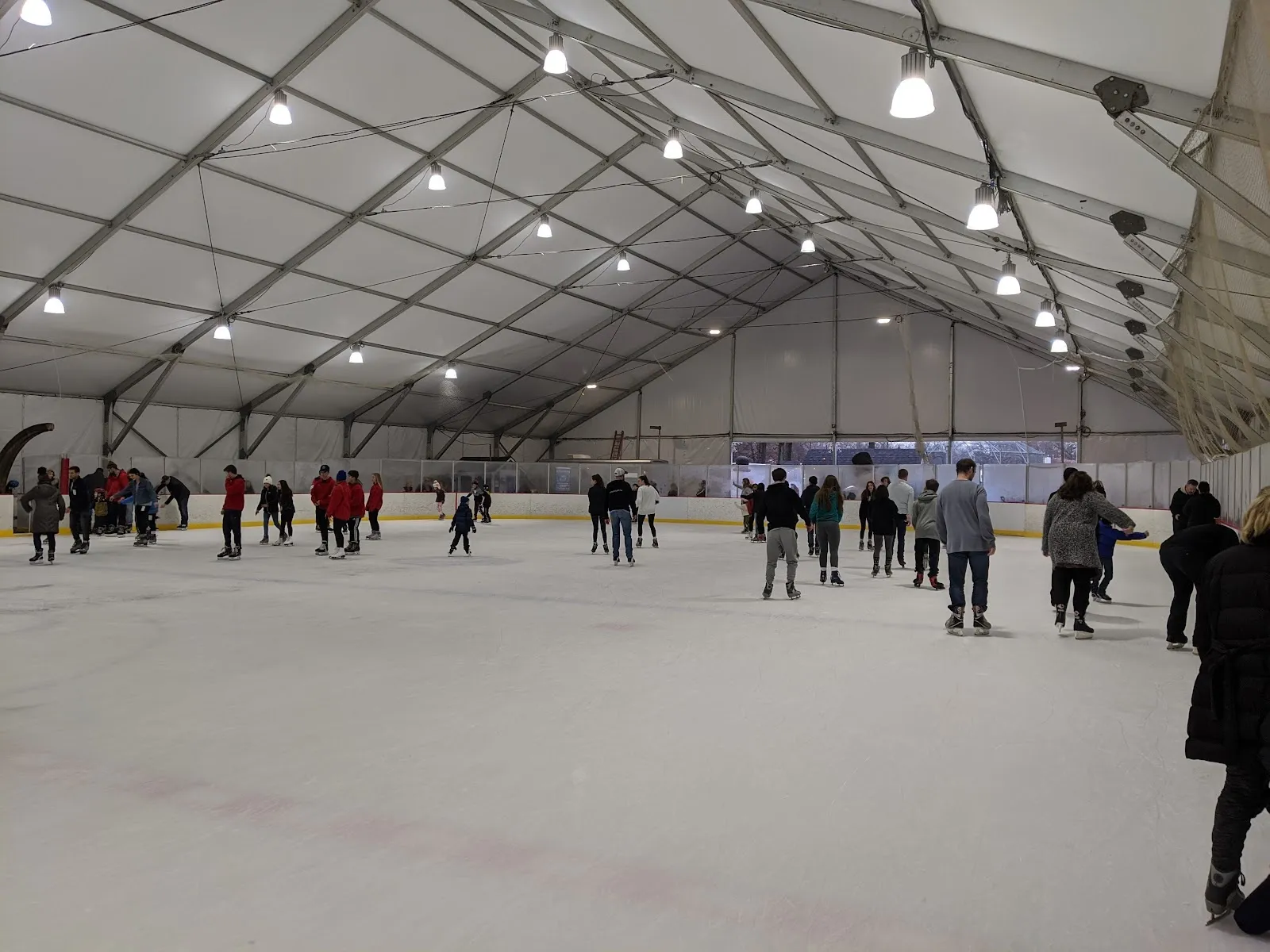Skaters spread across the ice inside a long white tent, steel ribs and lights overhead and a rink board along the far side. — The Westfield Rink
