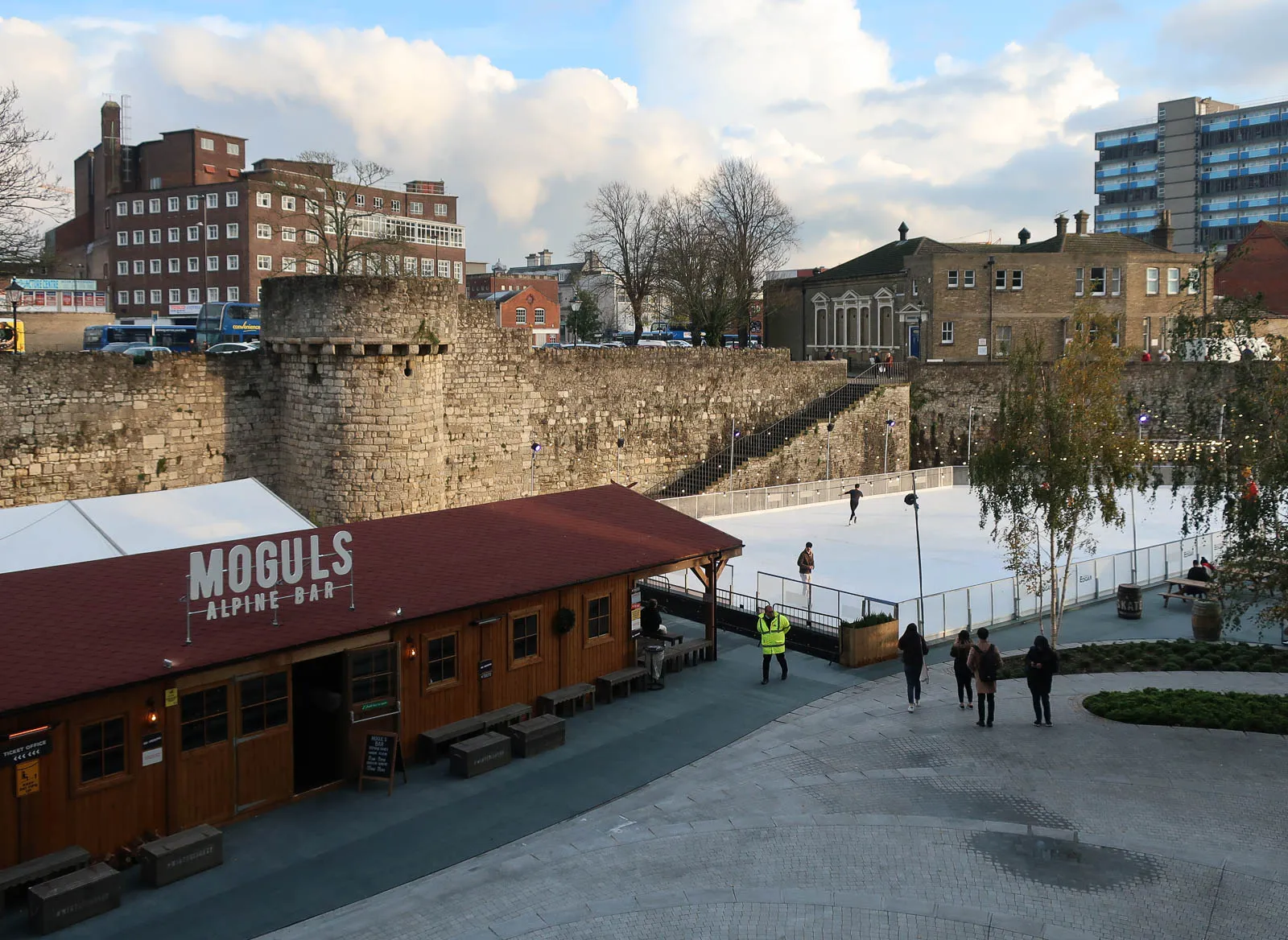 The outdoor ice rink beside Westquay in Southampton, with the medieval town wall behind it and a wooden alpine bar on the near side. — Westquay On Ice