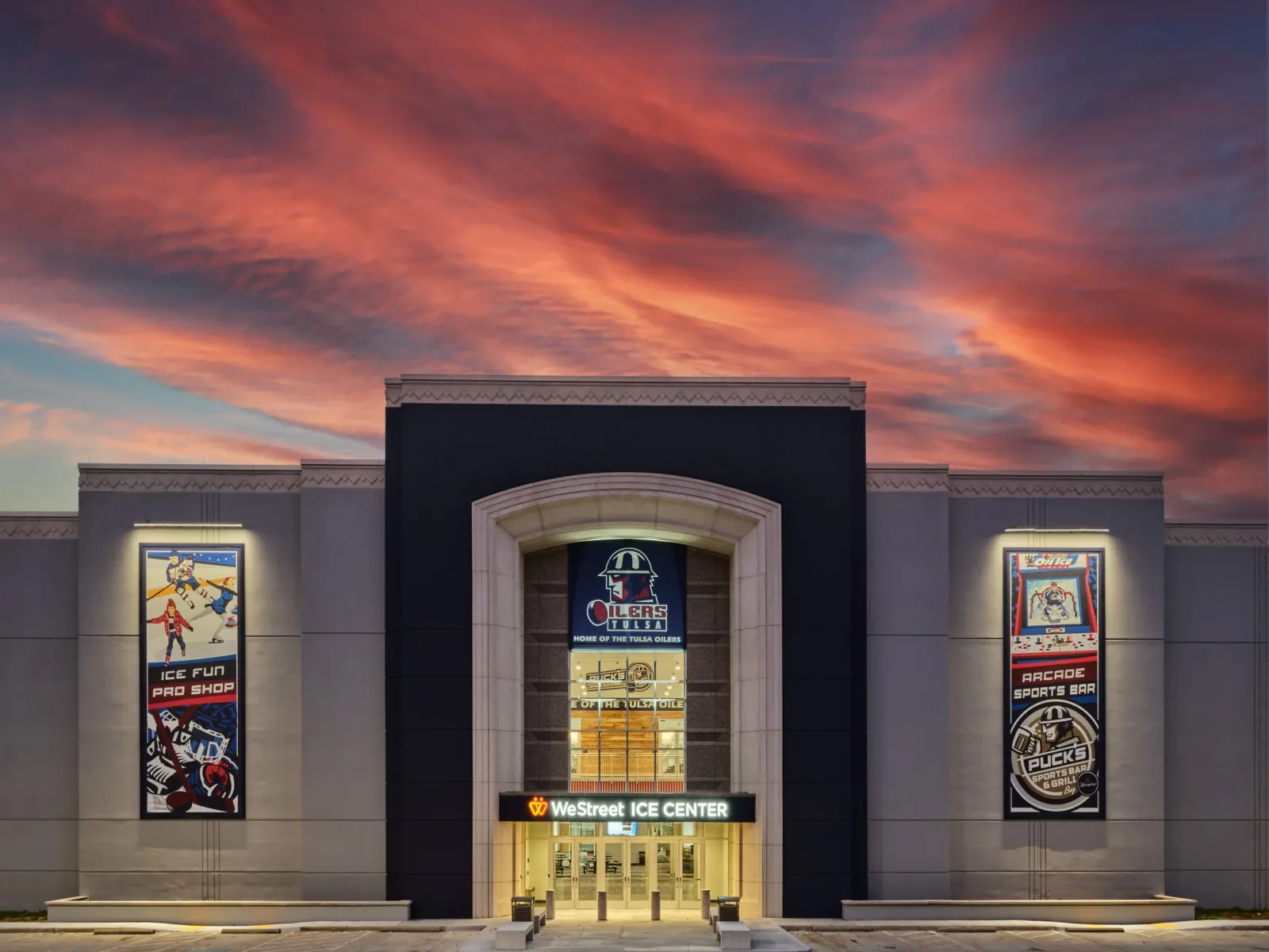 The WeStreet Ice Center entrance at dusk, with the Tulsa Oilers logo banner over the doors and Ice Fun Pro Shop and Arcade Sports Bar wall graphics on either side. — WeStreet Ice Center