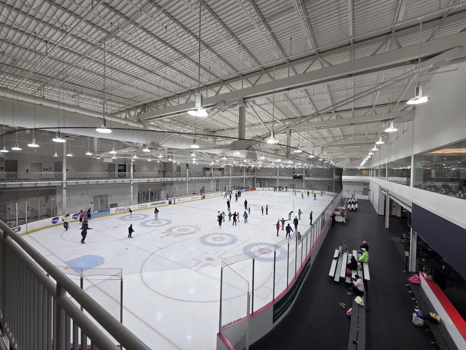 Elevated view from the mezzanine of the full ice sheet with curling circles painted in, dasher boards, and skaters spread across the surface during a public session. — WeStreet Ice Center