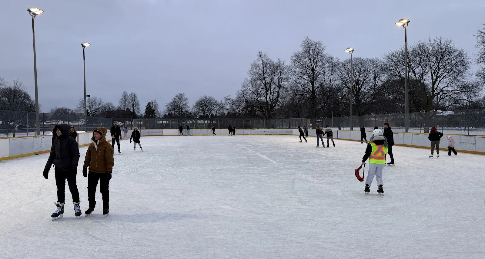 Wide view of the Westway outdoor rink with skaters spread across the ice at dusk and floodlights beyond the boards — Westway Park Outdoor Ice Rink