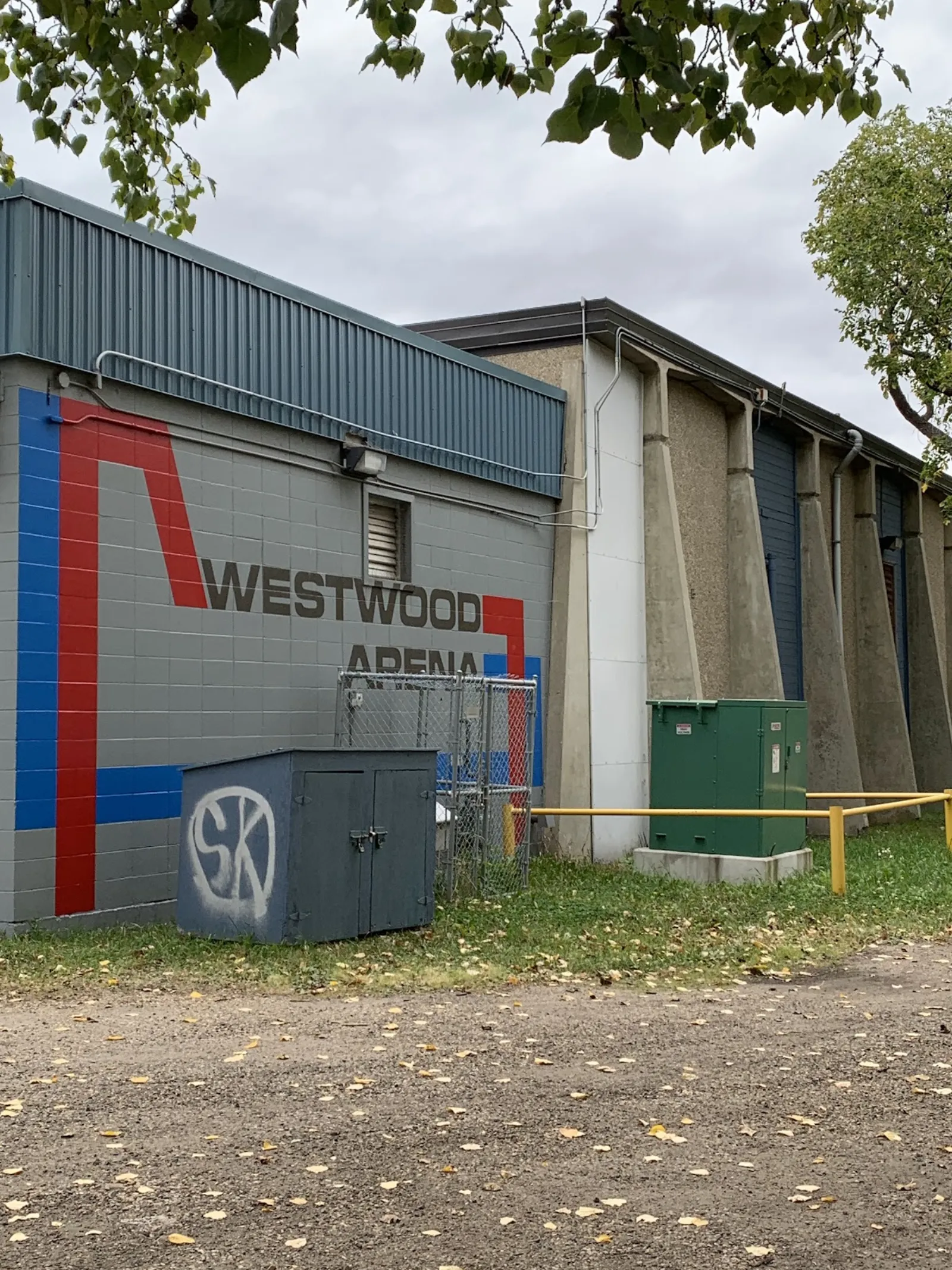 Costado del edificio gris de bloques pintado con un gráfico de franjas rojas y azules y las letras "WESTWOOD ARENA", con contrafuertes de concreto y puertas de carga a lo largo del muro, y cajas de servicios y un camino de grava al frente. — Westwood Arena