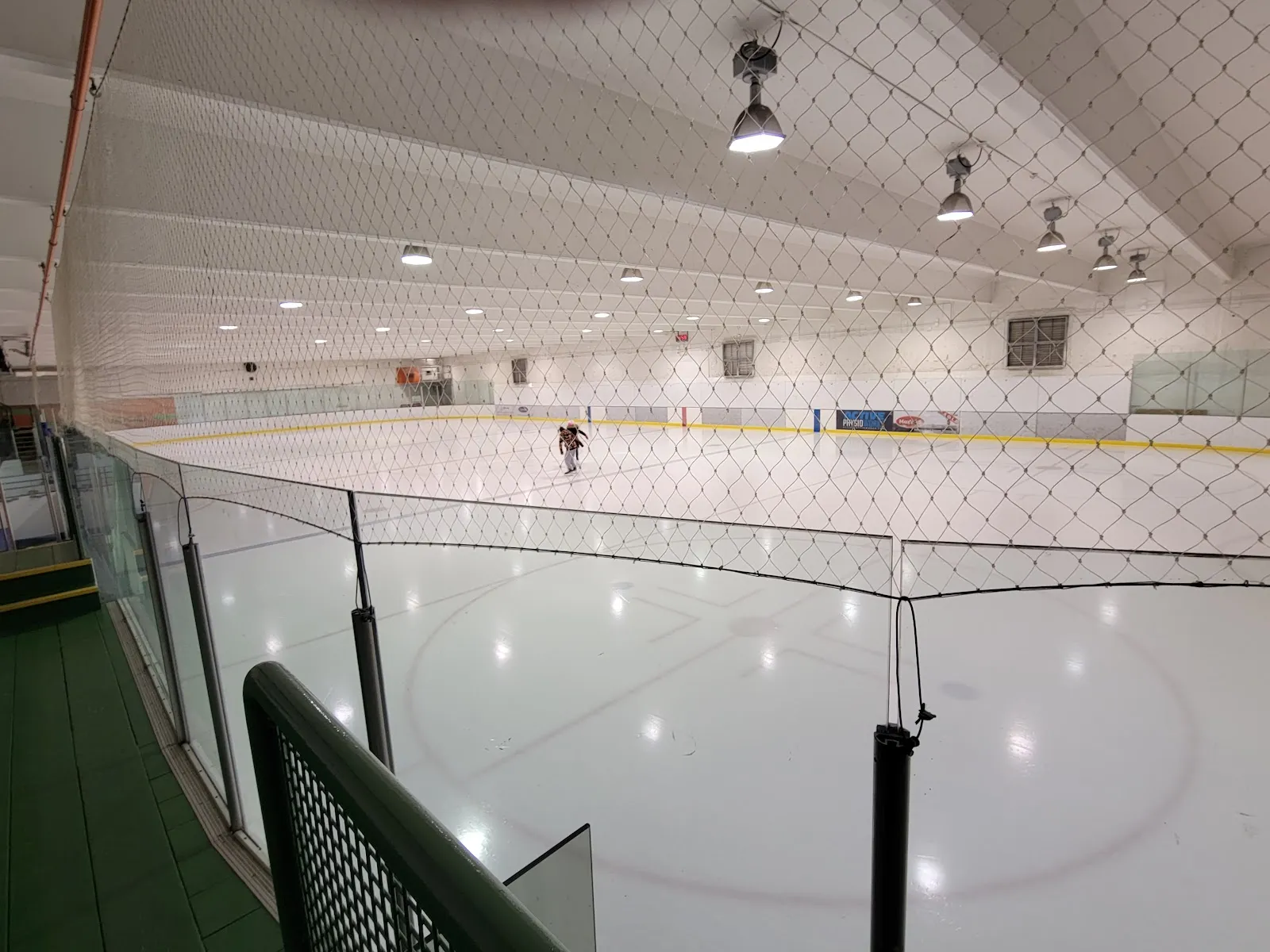 La pista vista desde un pasillo del nivel superior detrás de la red protectora, con toda la pista, vallas con anuncios de patrocinadores y un solo patinador a lo lejos cerca del centro de la pista. — Westwood Arena