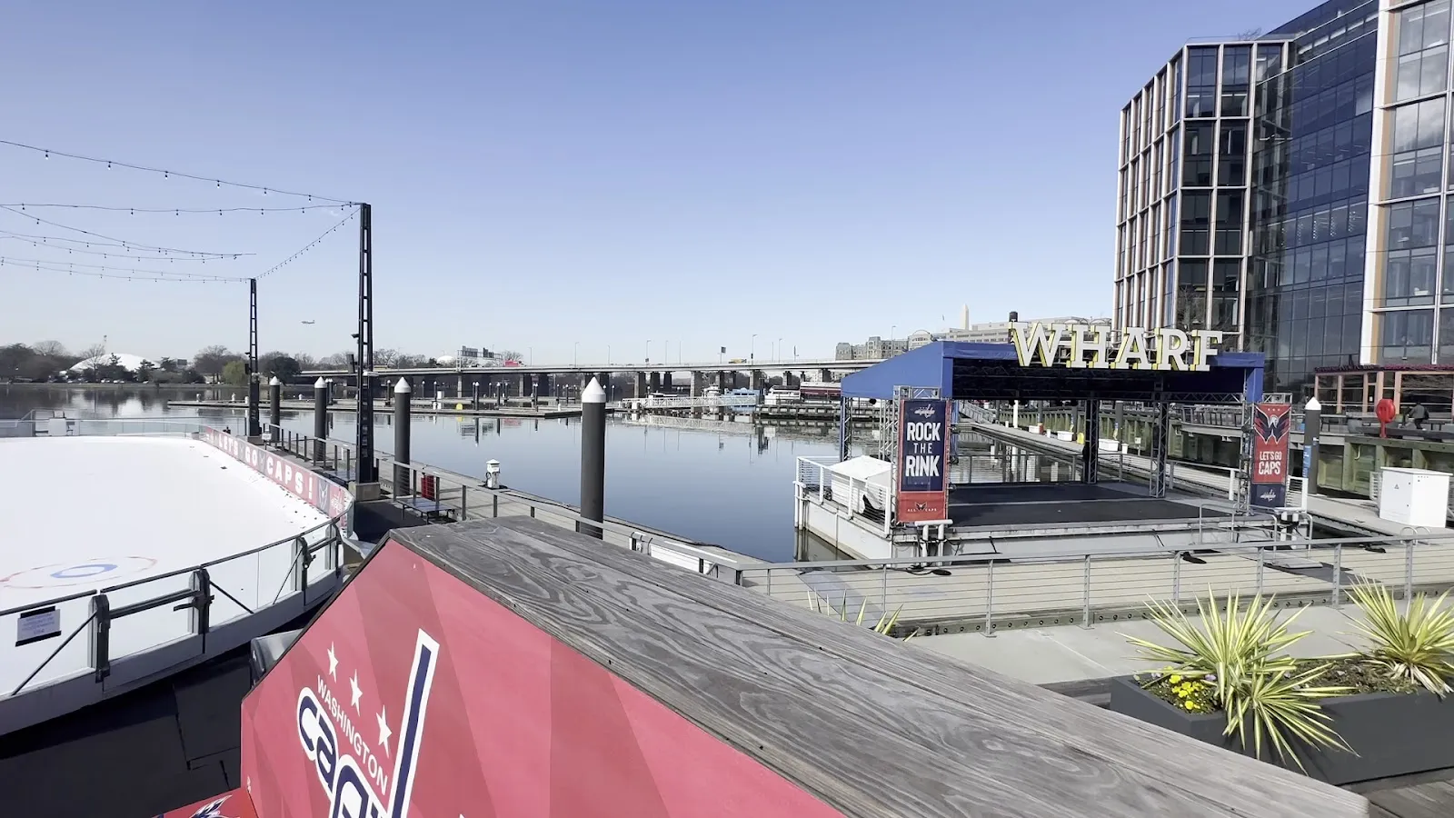 La pista del muelle de día, junto al letrero de The Wharf y el escenario flotante, con bandas de los Capitals y un rincón de hielo vacío. — The Wharf Ice Rink
