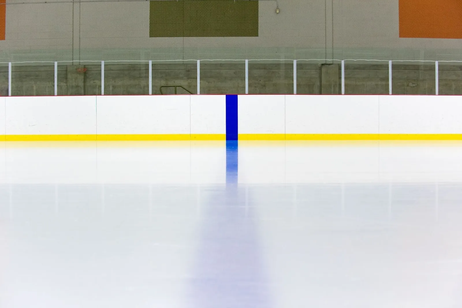 Close view from ice level of a hockey rink's white dasher boards with a yellow kick plate and a blue line painted on the ice, glass above the boards, and olive and orange colored wall padding with an exit sign visible behind. — Whatcom Sports and Recreation
