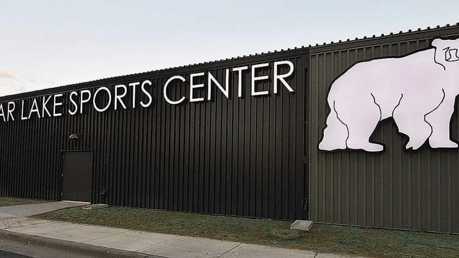 La pared exterior oscura revestida de metal con el nombre del Sports Center en letras grandes y un cartel de oso polar, vista desde la acera. — White Bear Lake Sports Center
