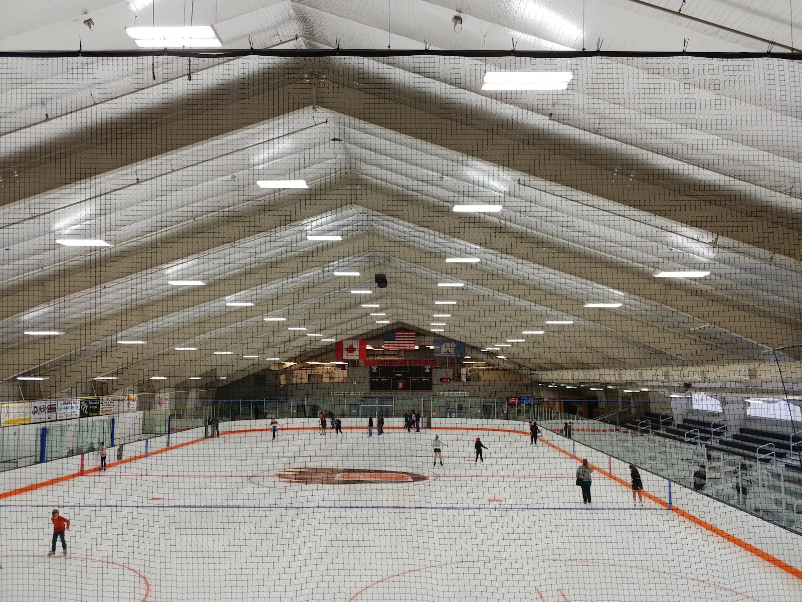 El hielo desde las gradas, con unos pocos patinadores, marcas naranjas y azules y las cerchas arqueadas de madera del techo. — White Bear Lake Sports Center