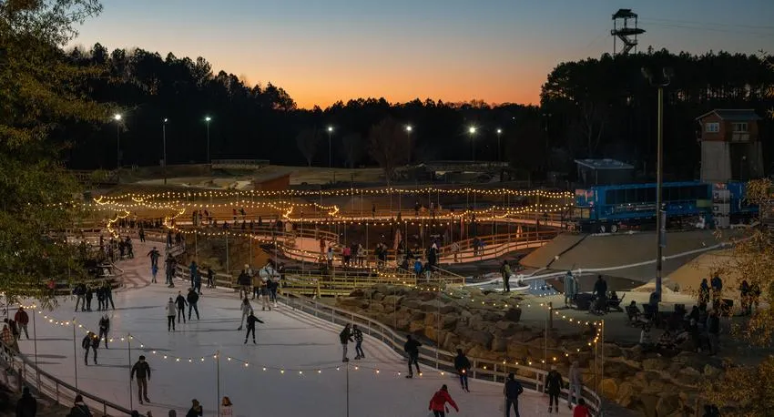 The outdoor rink at dusk from the bank above, lit by string lights, with the Whitewater Center's towers and trails behind it. — Ice Skating at the Whitewater Center