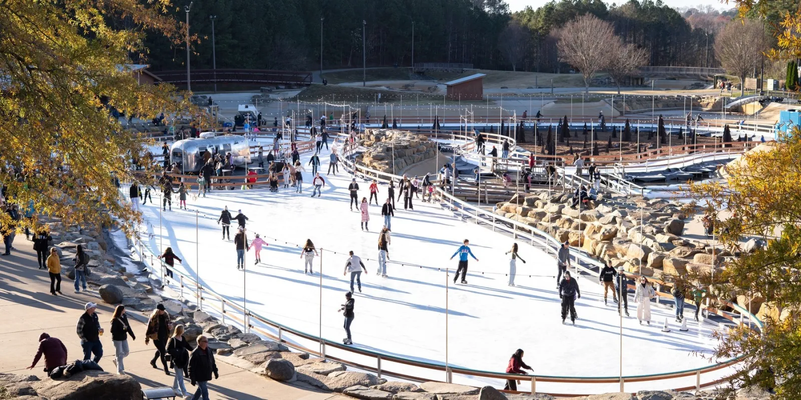 A wide view of the outdoor rink in the whitewater park, many skaters spread across the winding ice with rocks and bare trees around it. — Ice Skating at the Whitewater Center