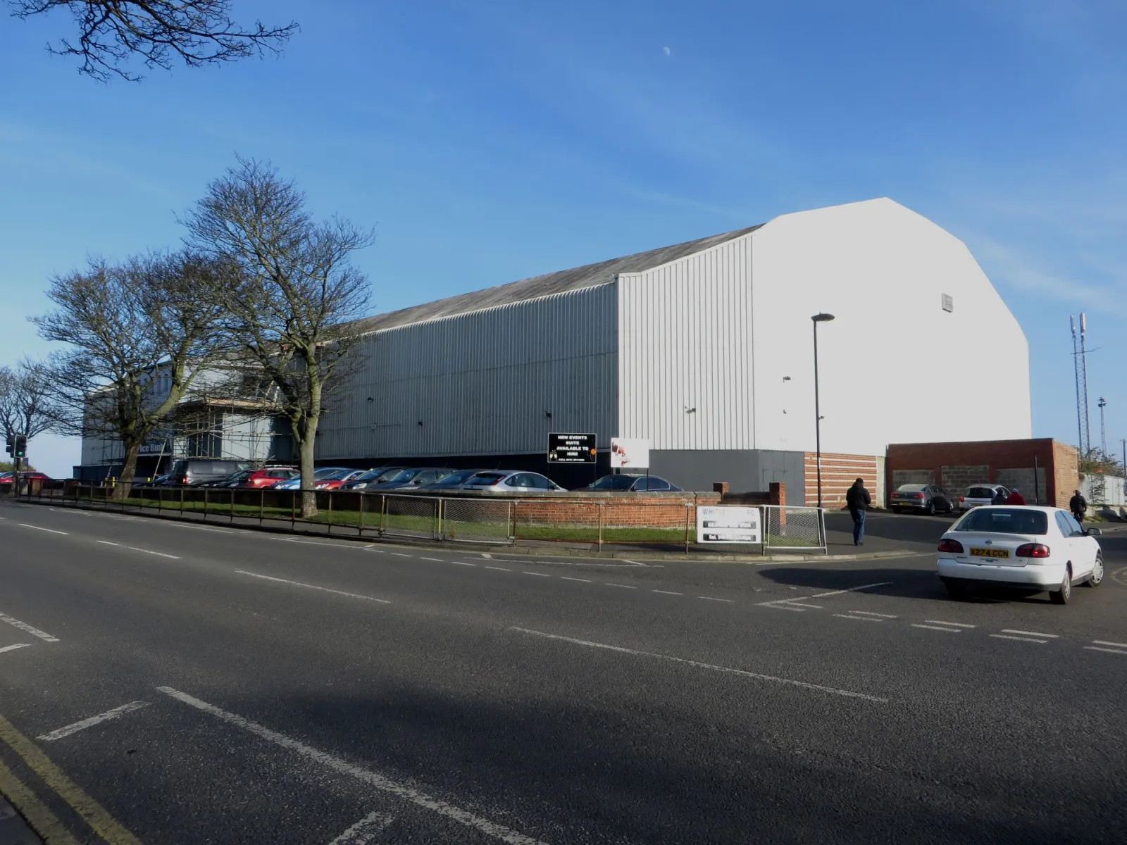 The whole Whitley Bay Ice Rink building seen across Hillheads Road on a clear day: a long grey clad hall with a tall white gable end. — Whitley Bay Ice Rink