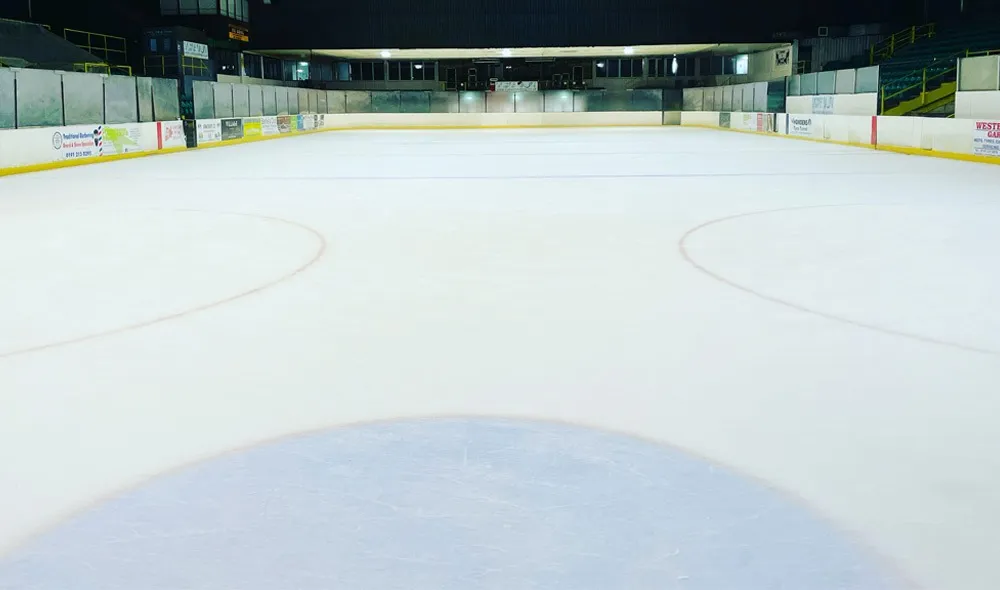 The empty ice surface at Whitley Bay with a face-off circle, the blue line and the boards and stands in the background. — Whitley Bay Ice Rink