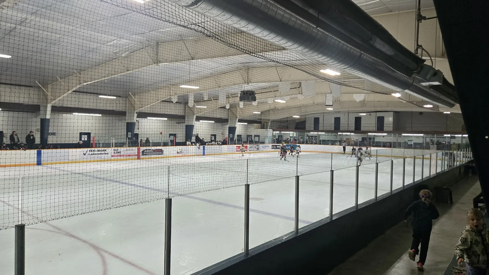 The ice at Whitney Arena seen from behind the boards netting during a youth game, with team banners hanging from the arched roof. — Whitney Arena