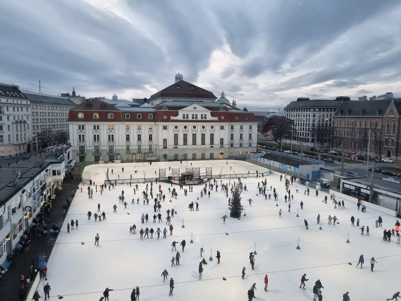 Die Eisbahn von oben in der winterlichen Dämmerung, sehr viele Läufer auf dem weißen Eis vor dem cremefarben-roten Konzerthaus. – Wiener Eislauf-Verein