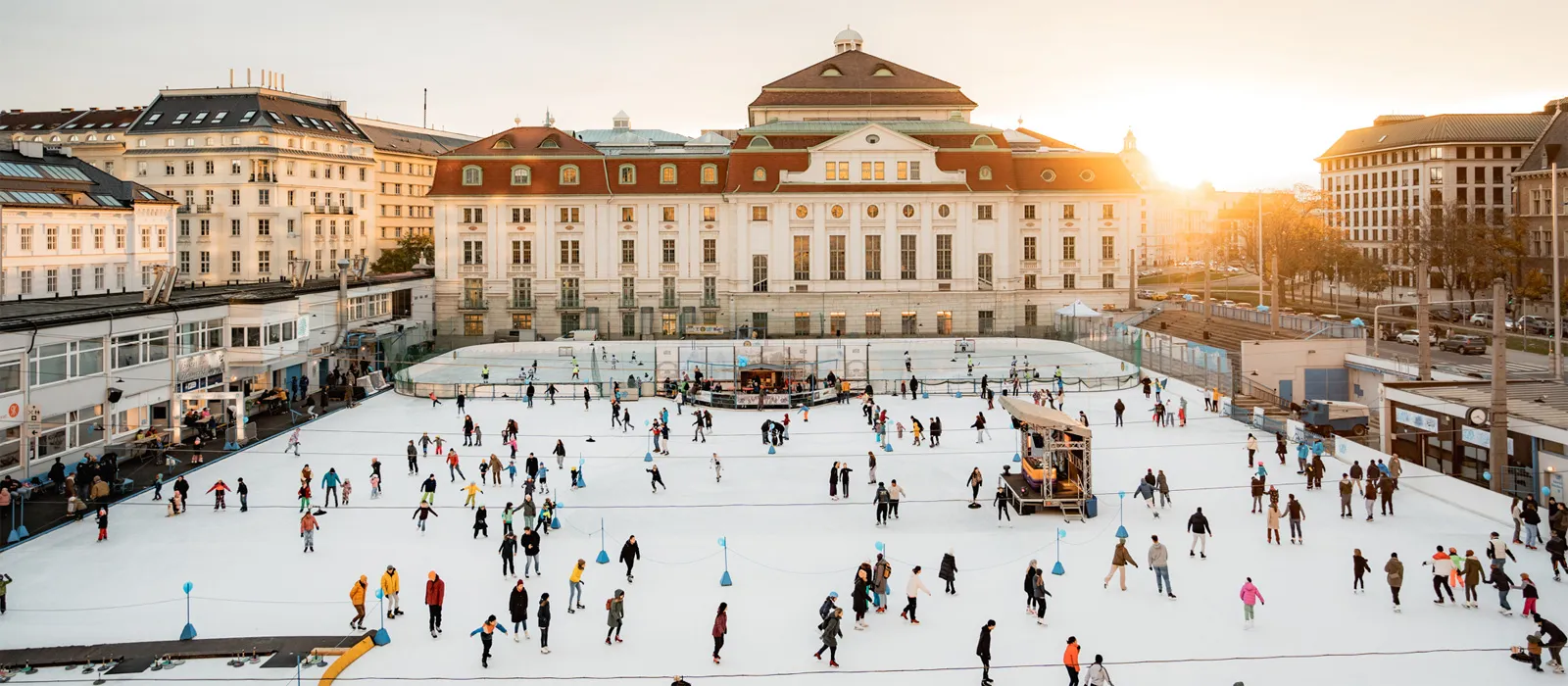Die gesamte Freiluft-Eisfläche des Wiener Eislauf-Vereins bei Sonnenuntergang, mit über die Fläche verteilten Läufern und dem Konzerthaus dahinter. – Wiener Eislauf-Verein