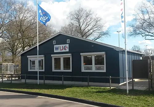 The dark-blue WIJC clubhouse beside the track, with the club flag, two ticket windows and the edge of the asphalt track in front. — Combibaan Wierden (Wierdense IJsclub)