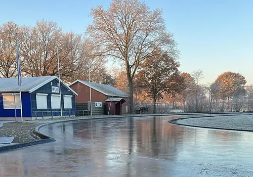 Frosted ice surface of the combi track at Sportpark Het Lageveld on a winter morning, with the blue clubhouse, bare trees and the curve of the roller track beside the ice. — Combibaan Wierden (Wierdense IJsclub)